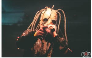 Capture d’écran 2026-10-15 à 20.43.11 Photo d’un chanteur masqué sur scène lors d’un concert de metal, portant un masque inquiétant orné de longues cordes et tenant un micro face au public. L’éclairage aux tons orangés contraste avec le fond sombre. Logo Rock’n’Hell visible en bas à droite.