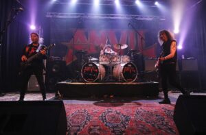 anvilmetalnews Le groupe de heavy metal Anvil en concert sur scène, avec le bassiste à gauche, le batteur au centre et le guitariste à droite, sous des éclairages violets et blancs.