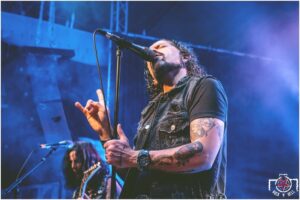 rocknhellronnie.jpg Chanteur de rock tatoué au micro sur scène, sous des lumières bleues et violettes, avec un guitariste en arrière-plan.