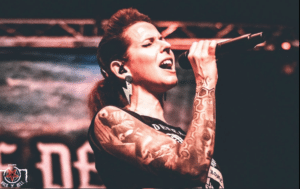 rocknhellwakethedead Photo d’une chanteuse tatouée en pleine performance sur scène, tenant un micro et chantant avec intensité. Elle porte un débardeur noir et des boucles d’oreilles en forme d’éclair, sous des éclairages aux tons rouges et orangés. Logo Rock’n’Hell visible en bas à gauche.