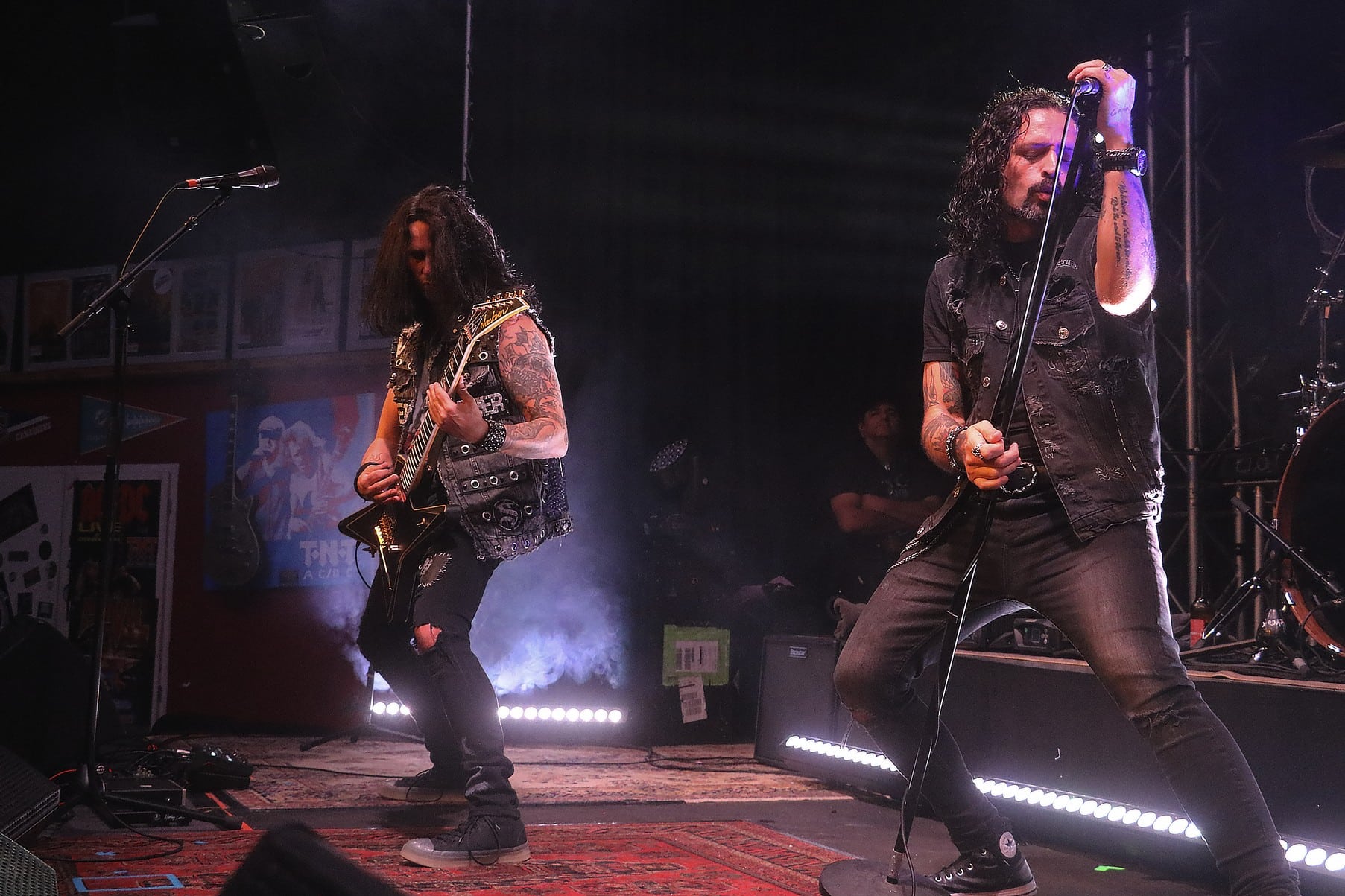 ronniemetalnews Groupe de hard rock en concert : chanteur tatoué au micro et guitariste aux cheveux longs jouant une guitare électrique, dans une ambiance sombre éclairée par des projecteurs et de la fumée.