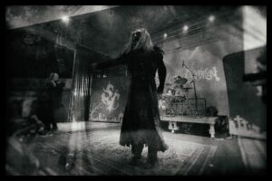 stcmetalnews Photographie en noir et blanc d’un concert de metal : un chanteur aux longs cheveux, vêtu d’un long manteau déchiré, se tient au centre d’une scène décorée de tombes et d’un crâne stylisé, tandis qu’un batteur joue en arrière-plan.